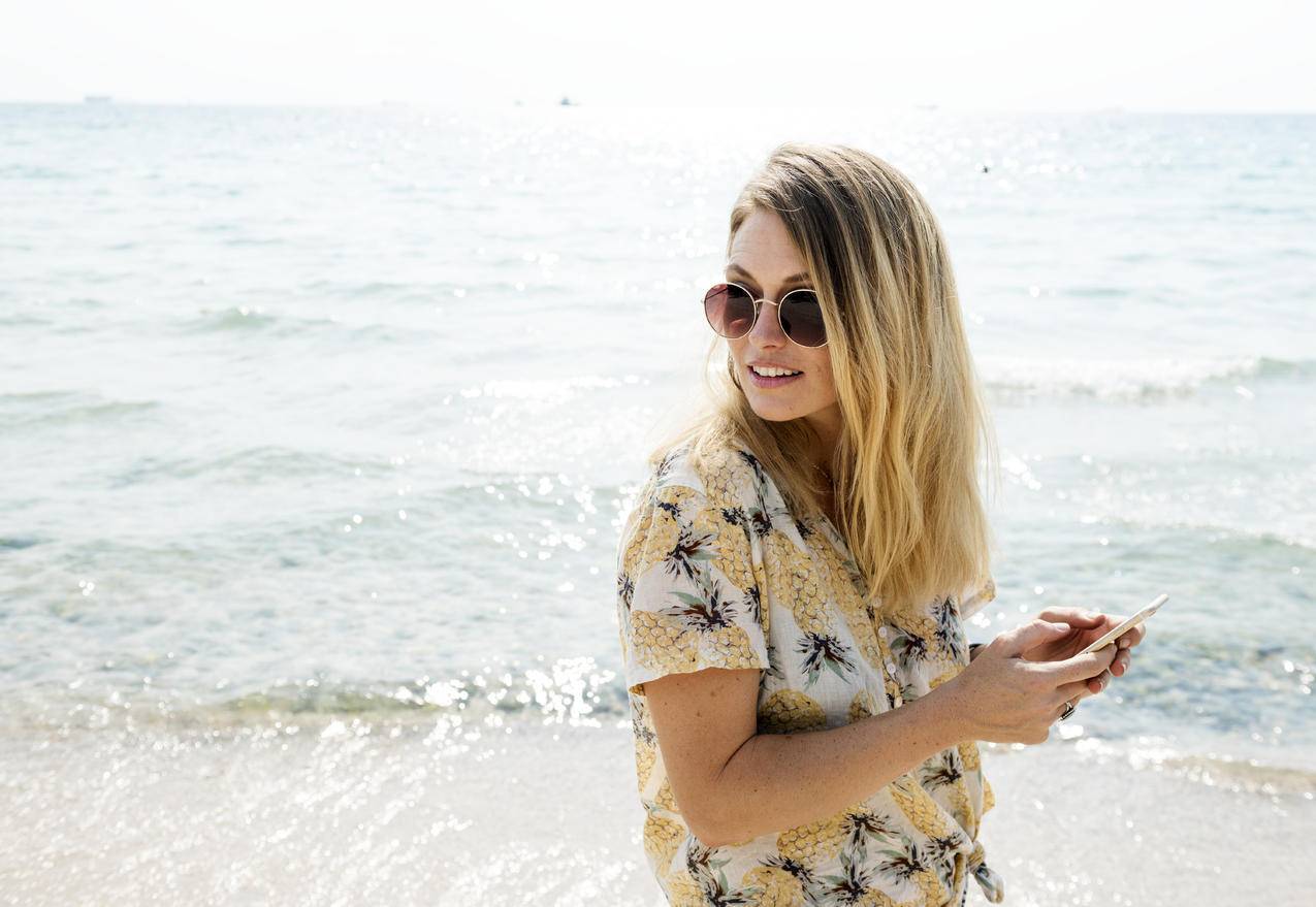 身穿黑色黄白相间钮扣衬衫的女子手持智能手机戴着太阳镜靠近水体