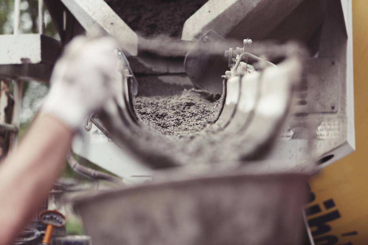 这张图片展示了一个建筑工地的景色在前景中,戴着工作手套的手拿着一个桶在后台可以看到水泥卡车水泥从卡车里流到桶里